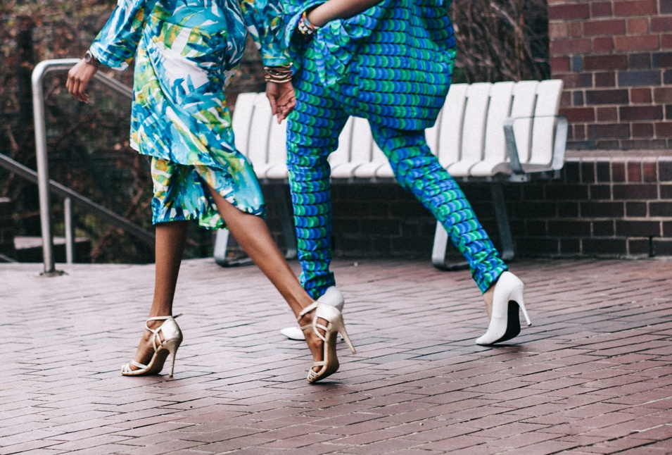 Cropped from the waist down, two women walking in high heel shoes.